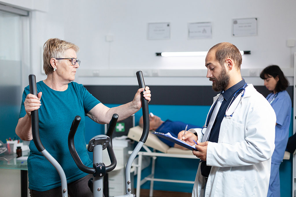 An Older Woman Using An Elliptical Machine While Talking With Her Physical Therapist Stages Of Cardiac Rehab