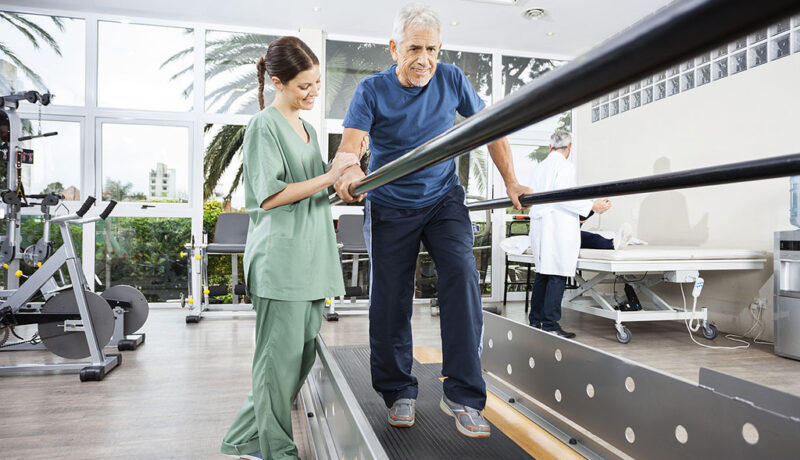 A Senior Man Walking Between Parallel Bars Being Helped By A Physical Therapist During Physical Therapy For Fall Prevention